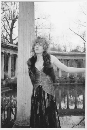 Monochrome image of a slender person with curly hair in a patterned vest and long skirt posing near a column in a park setting.