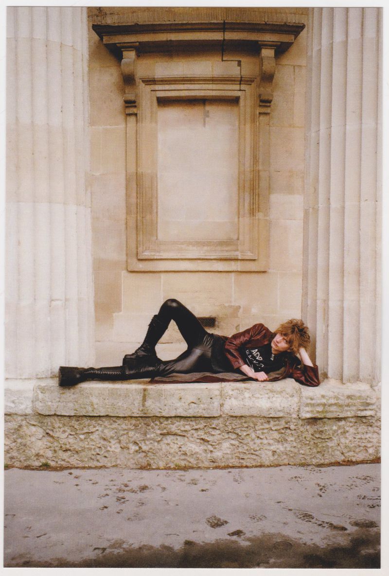 Adult with curly hair poses while lying on her side wearing boots and a brown jacket in front of an old stone column facade.