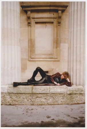Adult with curly hair poses while lying on her side wearing boots and a brown jacket in front of an old stone column facade.