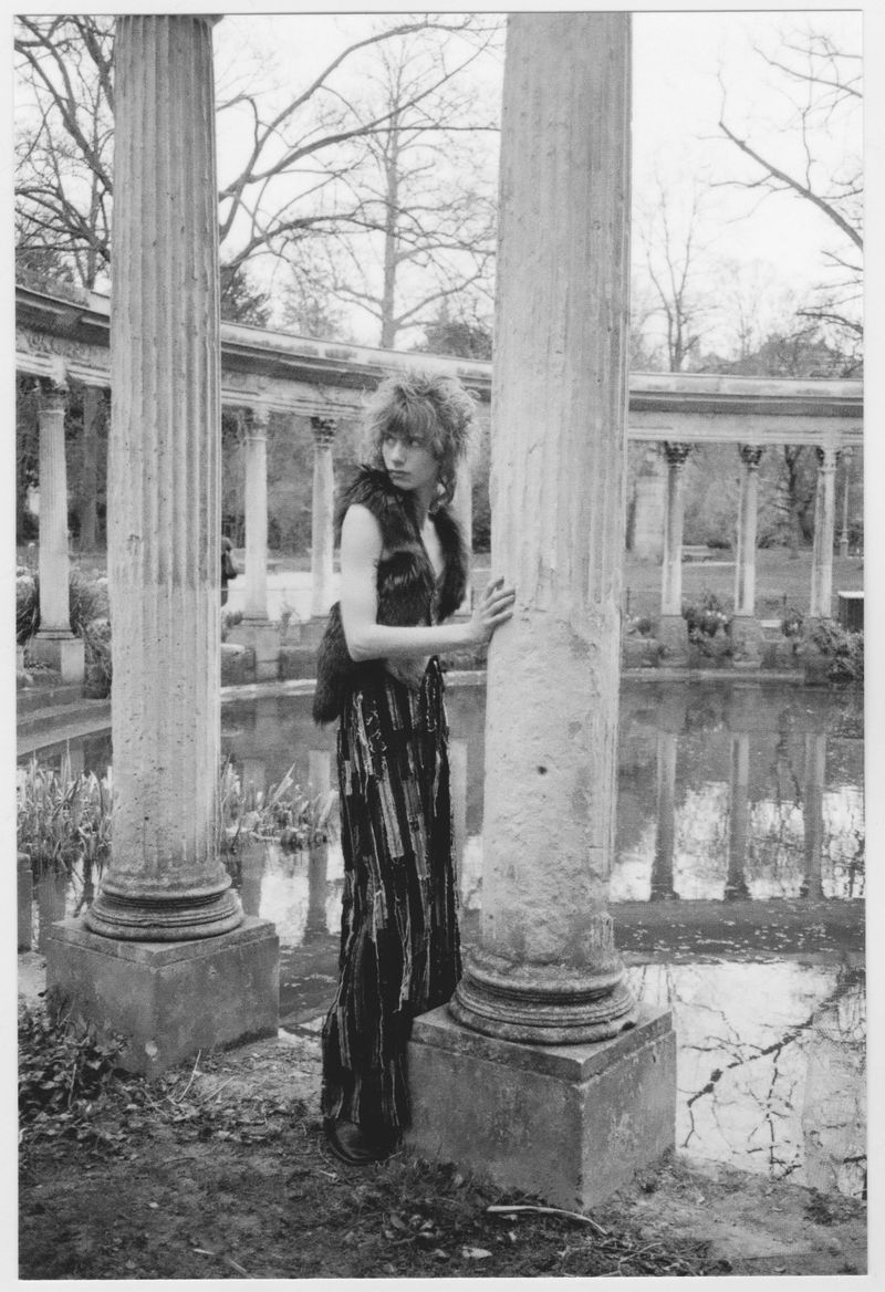 Adult woman poses in a fur vest and wide-leg pants next to a concrete column on a cloudy day, with water and architecture behind.