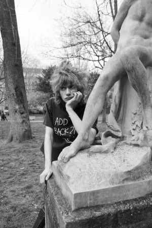 Monochrome shot of a young adult resting by a statue in a park. The image features a person in a t-shirt with text, and foliage backdrop.