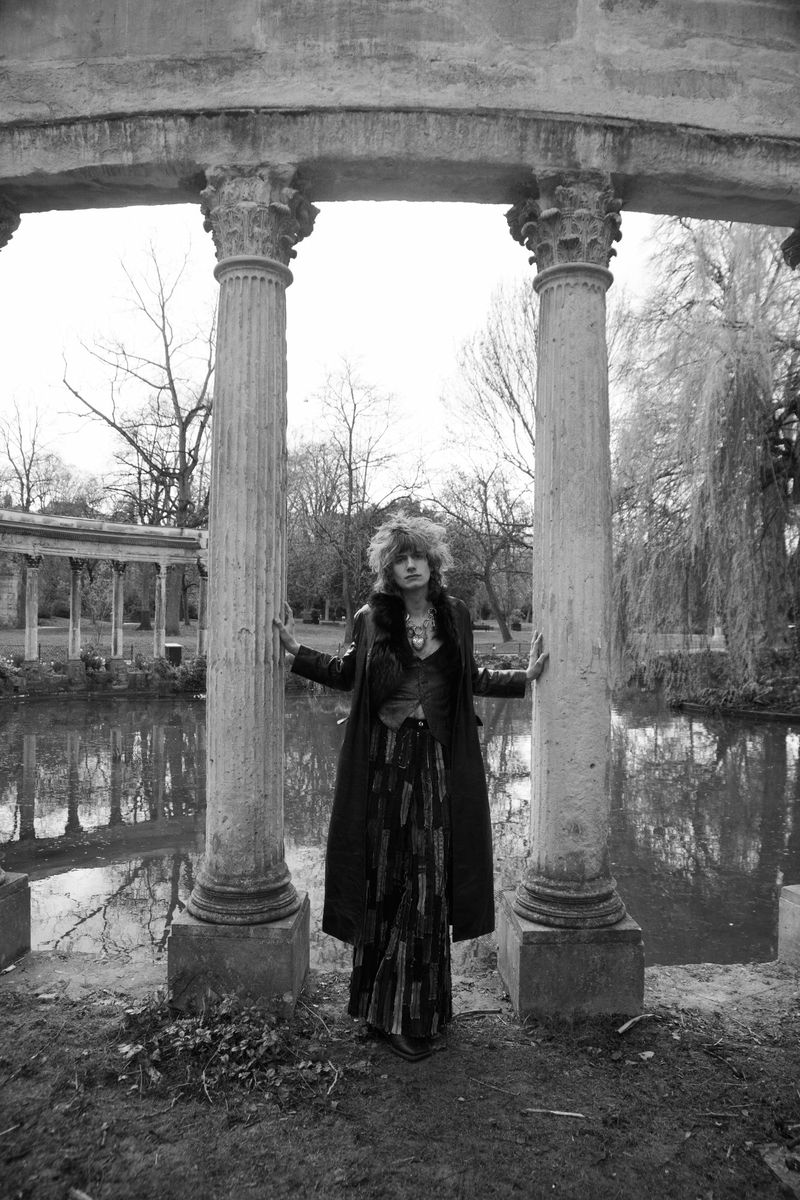 A woman with frizzy red hair in a coat and dress stands between stone columns near a pond, creating an editorial fashion shot outdoors.
