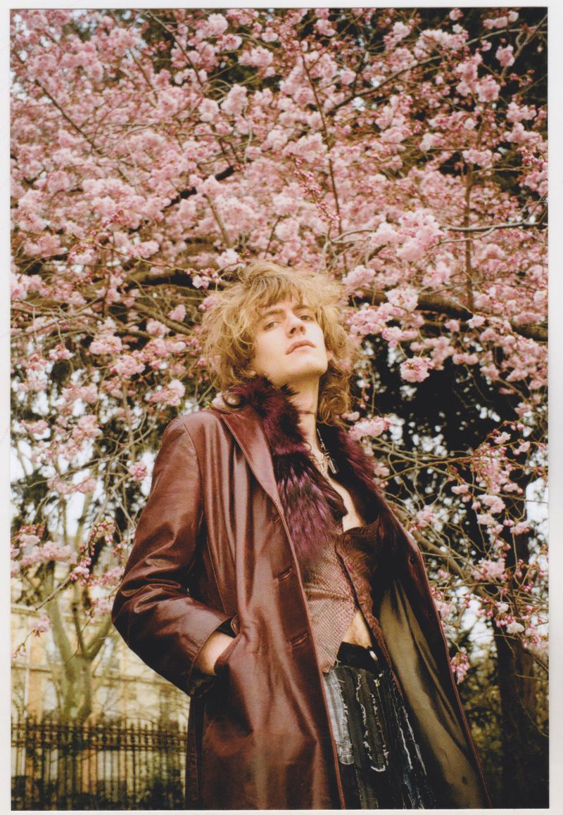 Adult male in a burgundy coat stands outdoors, under a tree with pink blossoms. He is gazing up at the sky. The lighting is soft.