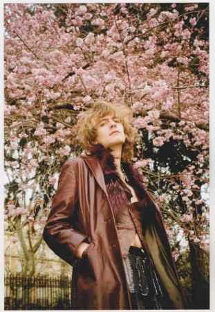 Adult male in a burgundy coat stands outdoors, under a tree with pink blossoms. He is gazing up at the sky. The lighting is soft.