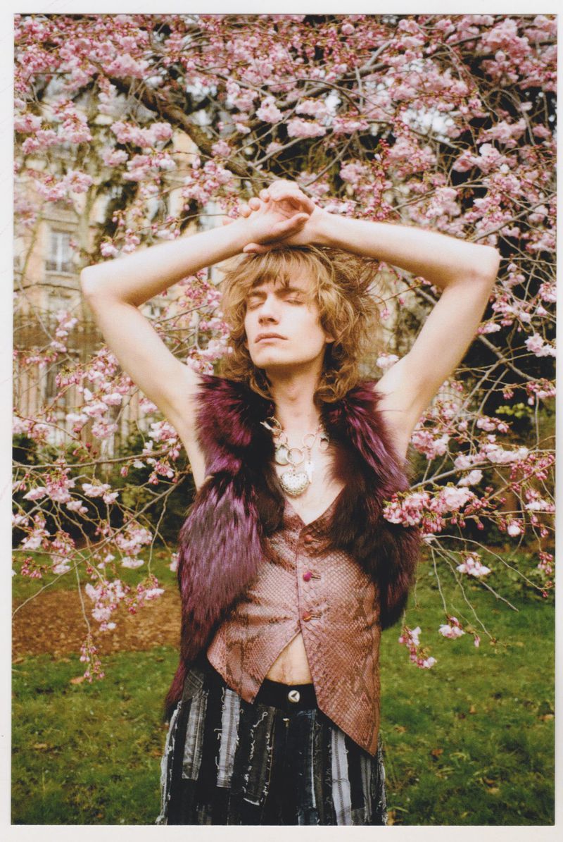 Young adult male poses outdoors in patterned vest and trousers, hands above his head against a background of pink blossoms and trees.
