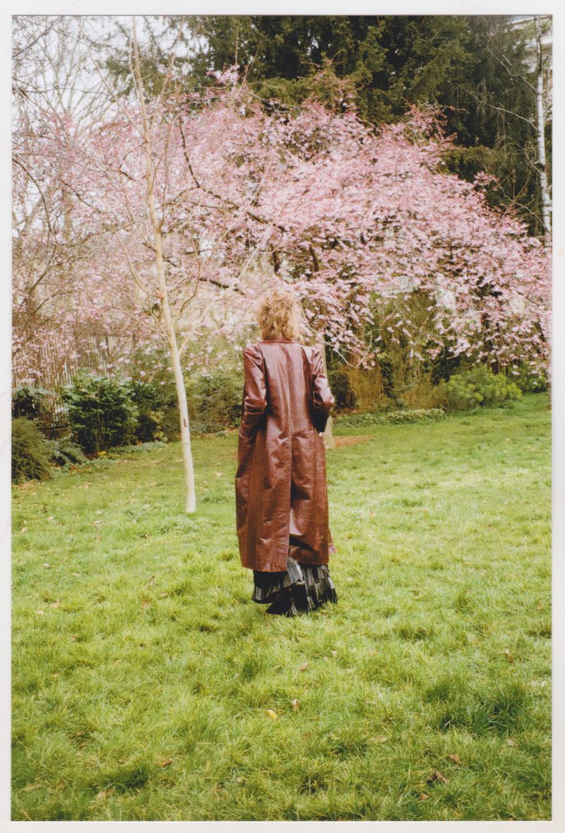 A woman wearing a brown coat stands in a grassy park with flowering trees, facing away from the camera on an overcast day.