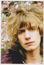 Portrait of a man with shaggy blonde hair and a dark patterned coat against a blurred background of pink flowers.