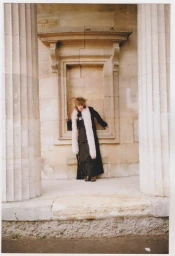 Model wearing a long black coat with a white fur stole standing between large neoclassical stone columns for Teeth Magazine