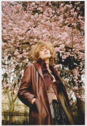 Model with long blonde hair wearing a brown leather coat standing beneath blooming pink cherry blossom trees