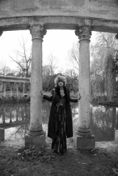 Model standing between classical stone columns by a body of water in a monochromatic outdoor fashion editorial image.