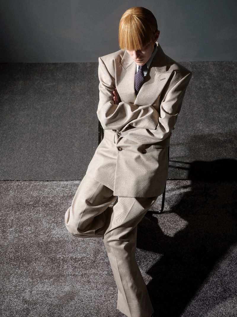 A young adult in a tan suit poses indoors, seated in a chair with arms crossed, wearing a tie and collared shirt, against a grey background.