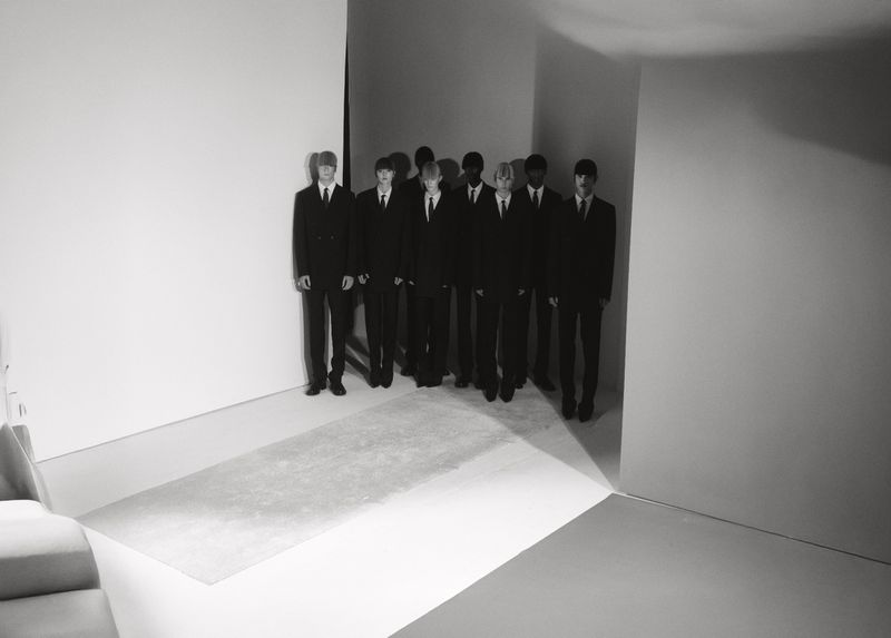 Group of formally dressed young men stand in a line against white walls, minimalist setting, monochrome, shadows, creating a stark visual statement.