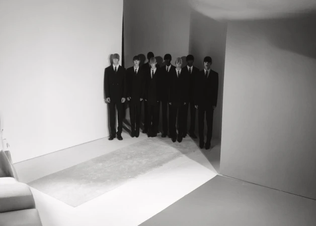 Group of formally dressed young men stand in a line against white walls, minimalist setting, monochrome, shadows, creating a stark visual statement.