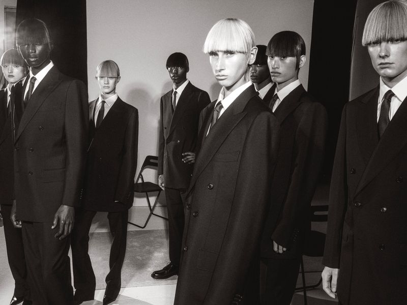 A group of androgynous fashion models with bowl haircuts, wearing suits and ties, stand in a photo studio with a white backdrop.
