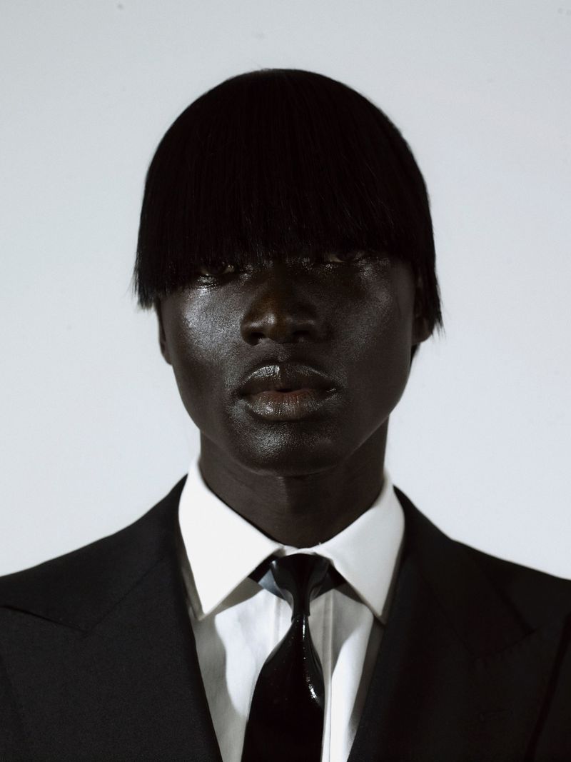 Close-up portrait of a black man wearing a black suit jacket, white shirt, and dark tie against a plain background