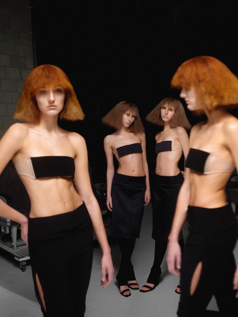 Four fashion models with angular auburn wigs pose in black tube tops and matching skirts against a dark background.