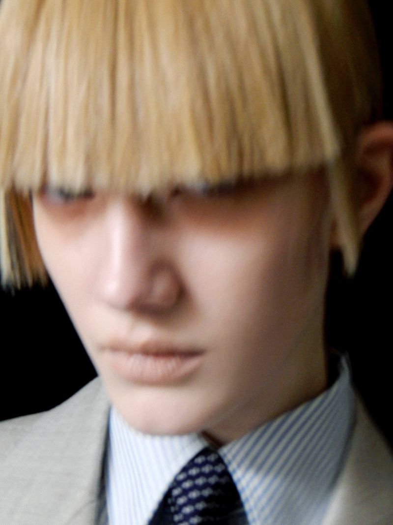 Close-up of a model with a blonde blunt bob haircut, wearing a light gray suit jacket, and striped collared shirt against a black backdrop.