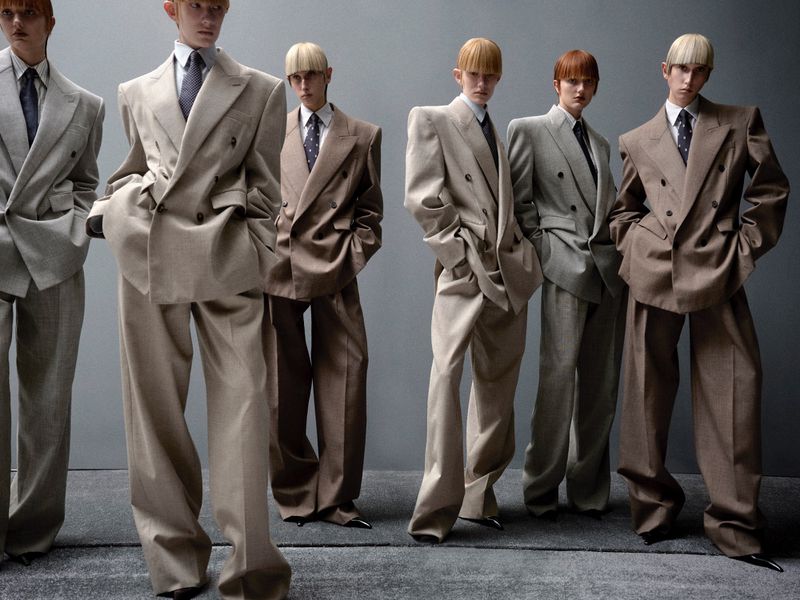 A group of androgynous fashion models wearing oversized suits posing in a studio with a gray backdrop, showcasing a modern, stylish aesthetic.
