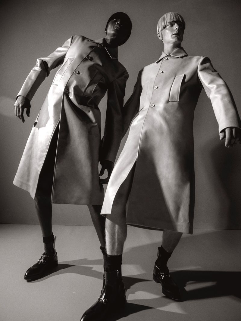 Two fashion models pose in matching trench coats and boots, low angle shot, studio lighting, black and white.