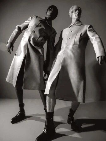 Two fashion models pose in matching trench coats and boots, low angle shot, studio lighting, black and white. 