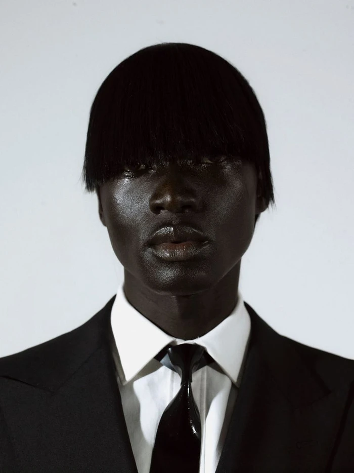 Close-up portrait of a black man wearing a black suit jacket, white shirt, and dark tie against a plain background