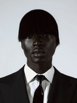 Close-up portrait of a black man wearing a black suit jacket, white shirt, and dark tie against a plain background