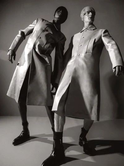 Two models wearing structured coats and boots posing against a plain studio background in a dramatic low-angle shot.