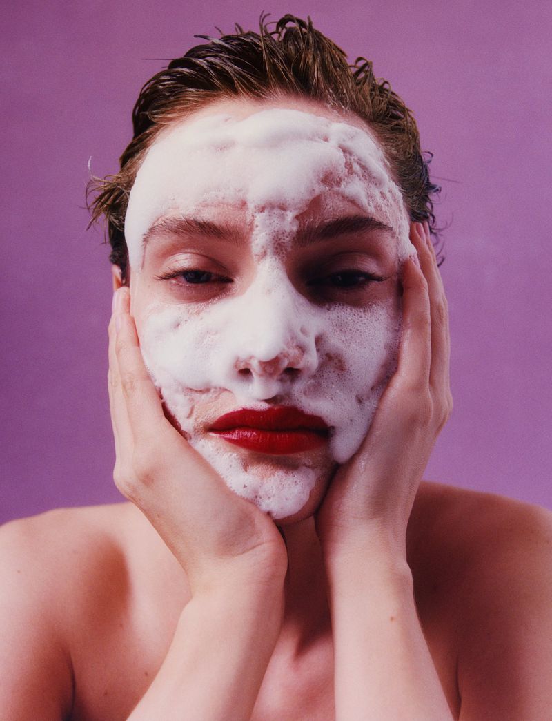 Close-up portrait of a model with white cleansing foam on her face and vibrant red lipstick against a mauve background.