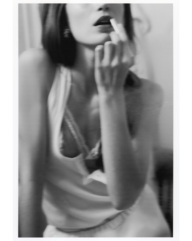 Monochrome portrait of an adult female applying lipstick, close-up, with blurred background, earrings, and an open tank top.