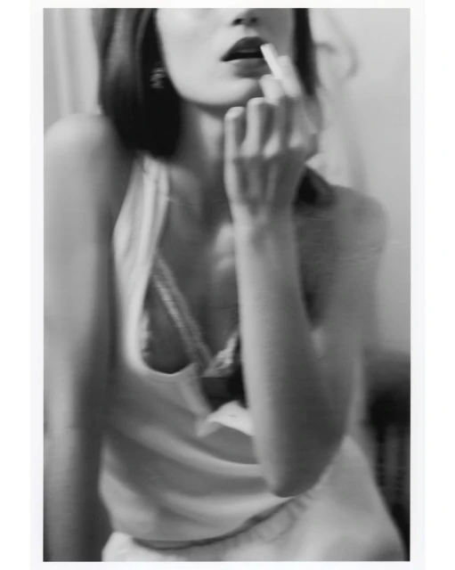Monochrome portrait of an adult female applying lipstick, close-up, with blurred background, earrings, and an open tank top.