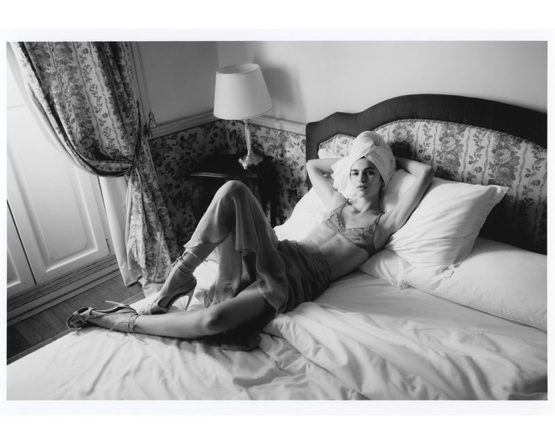 Black and white studio-style shot of woman reclining on a bed in lingerie with a towel wrapped around her head, looking at the camera.