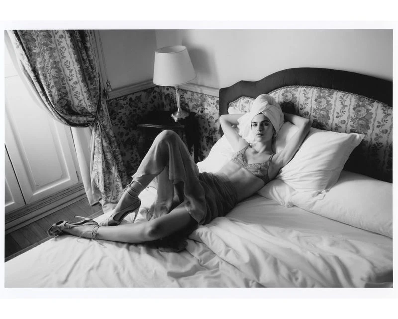 Black and white studio-style shot of woman reclining on a bed in lingerie with a towel wrapped around her head, looking at the camera.