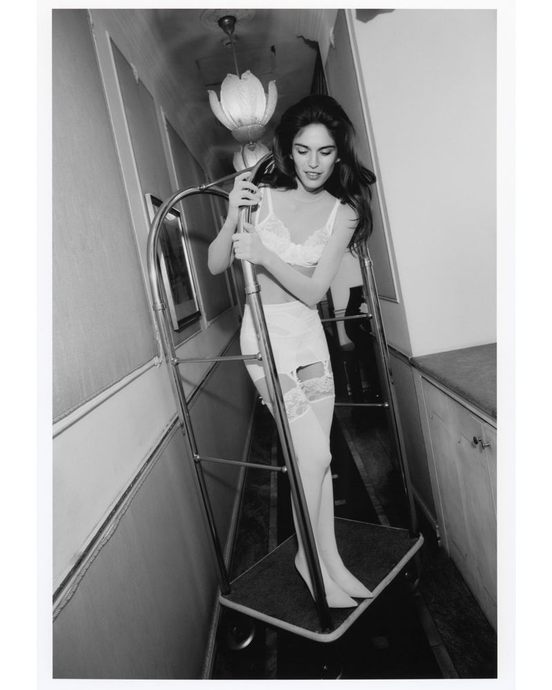 Black and white photograph of a woman in lingerie pushing a luggage cart in a hallway, with overhead lighting, in a monochromatic setting.