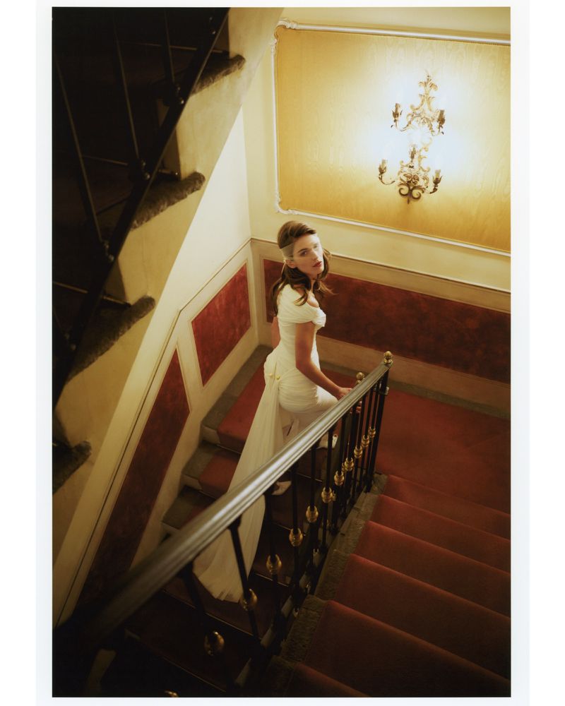 A woman in a white dress walks down a red-carpeted staircase in an interior, with a chandelier illuminating the scene.
