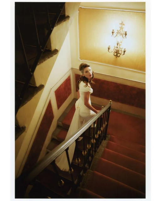 A woman in a white dress walks down a red-carpeted staircase in an interior, with a chandelier illuminating the scene.