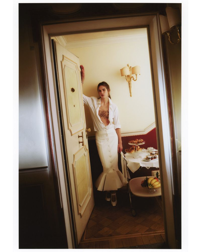 A woman poses in an open doorway wearing a white button-down shirt and white mermaid skirt, with a tea service visible in the background.