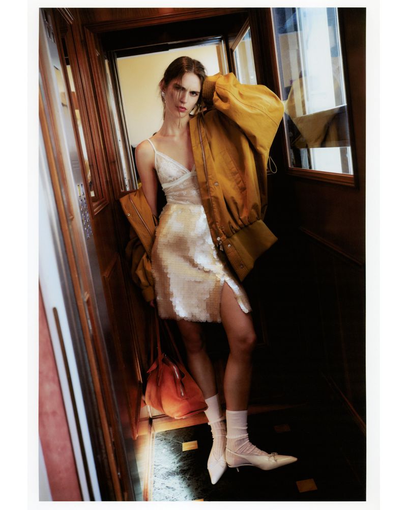 Fashion shot of a woman in a slip dress, wearing a jacket over her shoulder, with a purse and heels in an elevator.