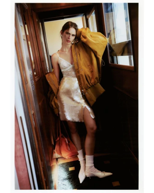 Fashion shot of a woman in a slip dress, wearing a jacket over her shoulder, with a purse and heels in an elevator.