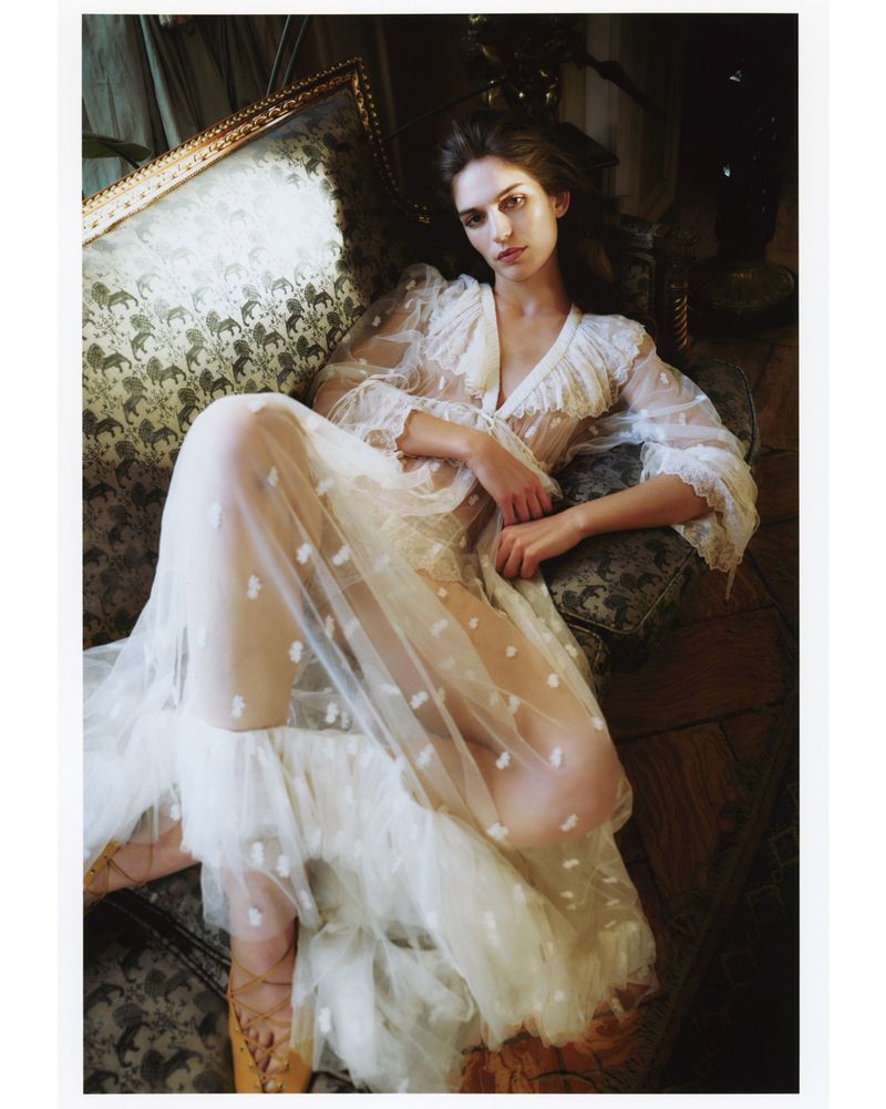 Woman in a flowing white dress lounges on an ornate vintage sofa in a dimly lit room, creating a dreamy, romantic fashion aesthetic.