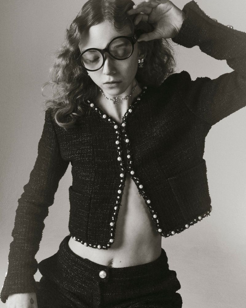 Woman in tweed suit and glasses poses in a studio. The model is wearing a pearl-trimmed tweed jacket and matching trousers.