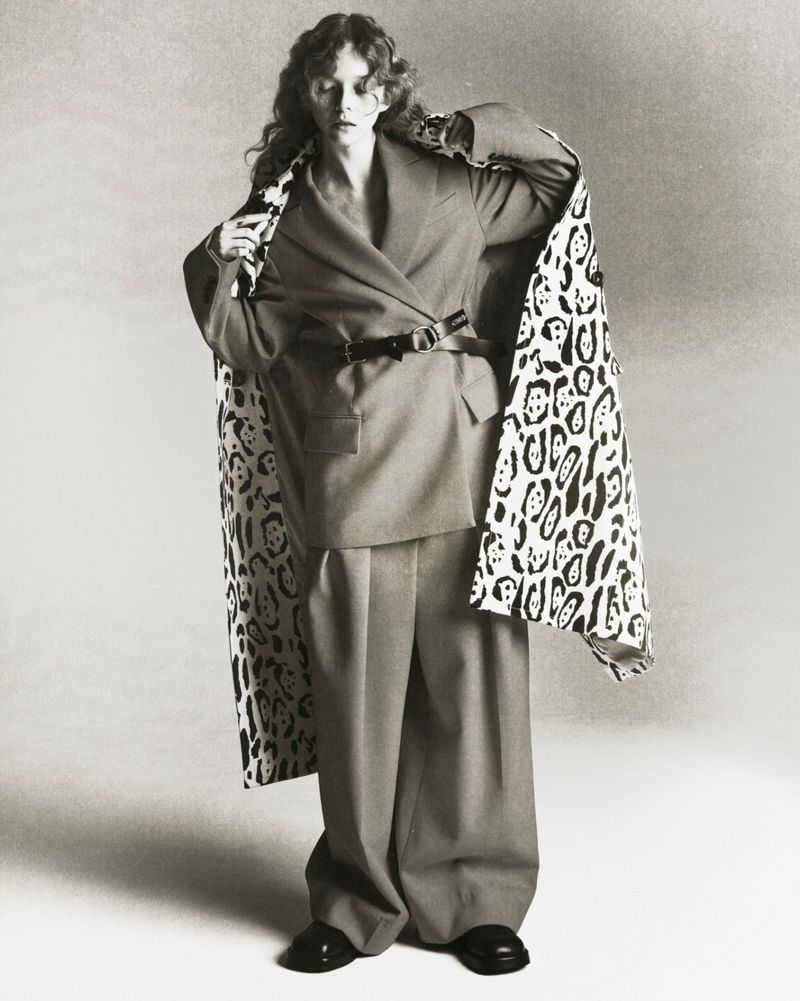 Woman in a grey suit with wide-leg pants and a black belt, draped with a black and white printed shawl, stands in a studio setting.