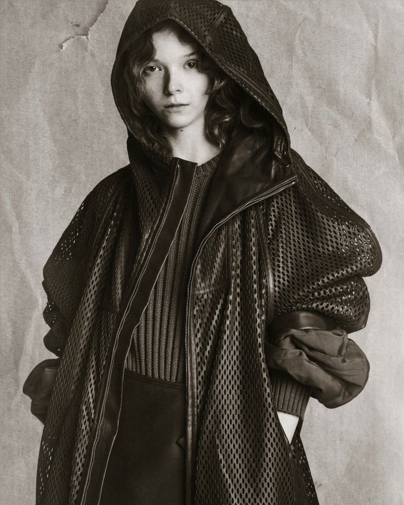 A young woman in a black hooded mesh coat poses against a mottled grey backdrop, showcasing a minimalist and slightly edgy fashion style.