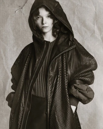 A young woman in a black hooded mesh coat poses against a mottled grey backdrop, showcasing a minimalist and slightly edgy fashion style.