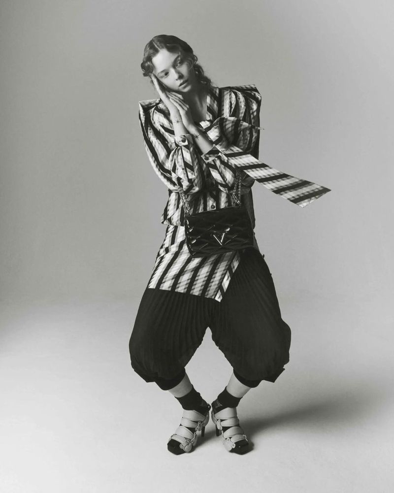Monochrome fashion shot of a young woman in a striped jacket, black pants, and platform shoes, posing against a gray backdrop.