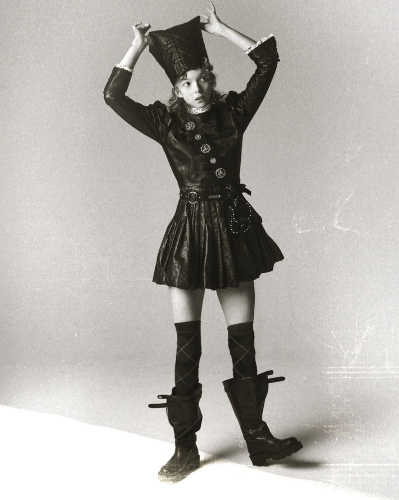 A young woman in a black outfit, hat, and boots poses in a full shot against a gradient gray background. Her hands are above her head.