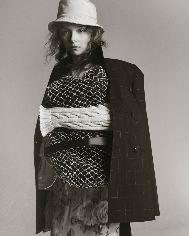 Model wearing a bucket hat, patterned sweater, and dark coat draped over shoulders against a neutral grey studio background.