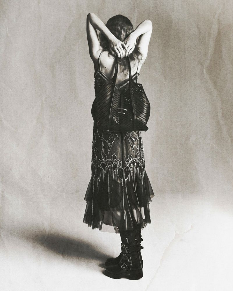 Back view of a woman wearing a dress and boots with a black bag over her shoulder, photographed in a studio setting.