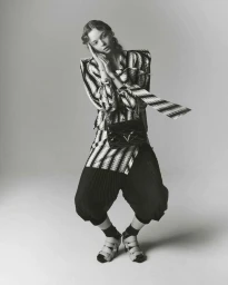 Model Lily Nova in striped top and dark trousers holding a black handbag against a grey seamless background.