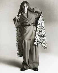 Model wearing an oversized suit and belted jacket draped over shoulders against a seamless studio backdrop in monochrome.