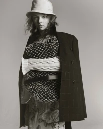 Model wearing a bucket hat, patterned sweater, and dark coat draped over shoulders against a neutral grey studio background.